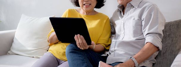 幸せなシニアカップルは,タブレットを使用してソファーに座って,自宅のリビングルームで家族とオンラインビデオ通話,楽しさとリラクゼーションで家族と遠くの幸せな老人と女性の会話. - 写真・画像