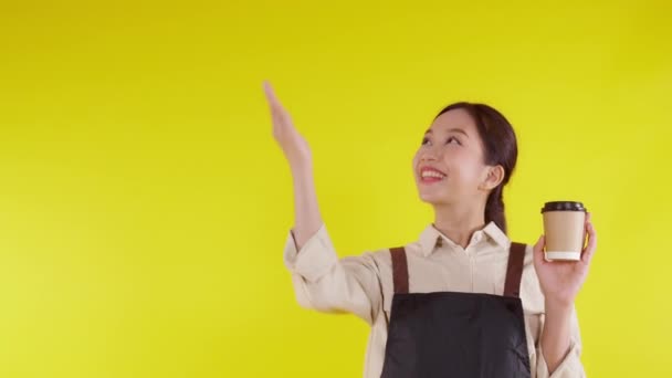 Retrato de una joven barista asiática usando delantal sosteniendo taza de café y presentando sobre fondo amarillo, camarera o emprendedora alegre y mostrando, pequeña empresa o startup, camarera de cafetería. - Metraje, vídeo