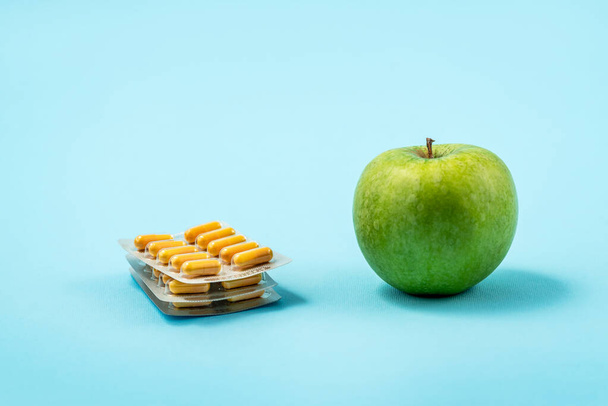 Prášky kapsle léků izolované na modrém pozadí versus zelené jablko. Koncepto léků a zdraví - Fotografie, Obrázek