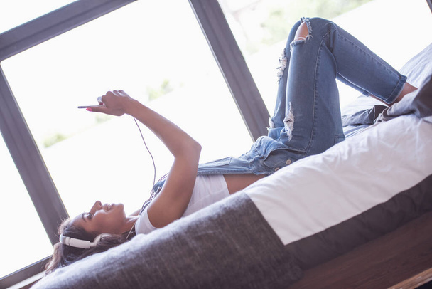 Mooi meisje in denim overall en hoofdtelefoon is het luisteren naar muziek met behulp van een smartphone en glimlachen terwijl liggend op bed thuis - Foto, afbeelding