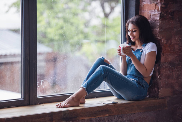 Menina bonita em geral denim está segurando uma xícara e sorrindo enquanto sentado no peitoril da janela em casa - Foto, Imagem