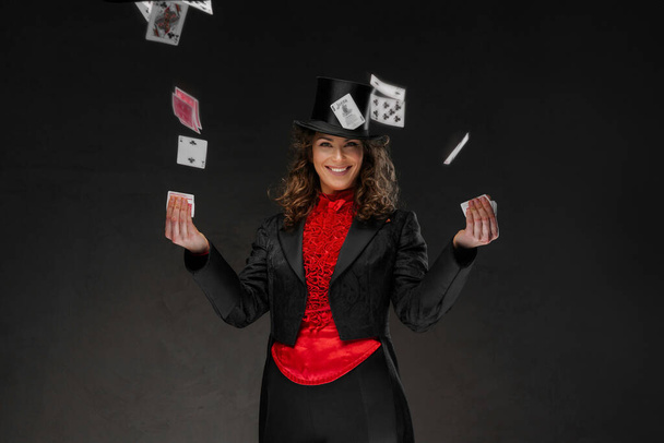 Очаровательная женщина-фокусница в костюме фокусника и черной цилиндре, выполняющая карточные трюки на тёмном фоне - Фото, изображение