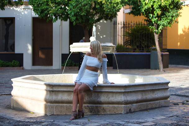 Hermosa joven rubia de los Estados Unidos está en un viaje de turismo en Sevilla, España. La chica es feliz y está sentada en una fuente de mármol. Concepto de viaje - Foto, imagen