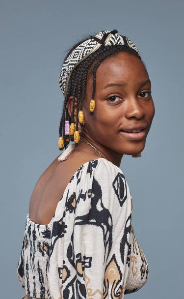 Портрет африканской девочки-подростка с африканской прической и национальной одеждой, смотрящей в камеру на синем фоне - Фото, изображение