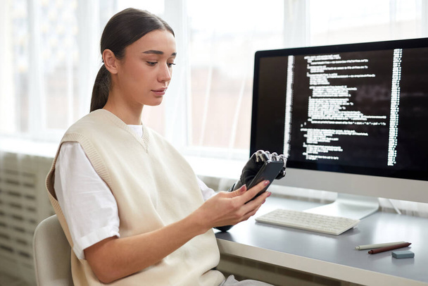 Jonge vrouw met een handicap met behulp van haar smartphone op het werk, terwijl zitten aan tafel met de computer - Foto, afbeelding