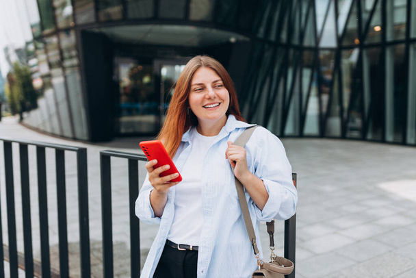 Νεαρή ευτυχισμένη επιχειρηματίας χαμογελά και χρησιμοποιεί έξυπνο τηλέφωνο στην πόλη. Όμορφη γυναίκα στο δρόμο για το γραφείο ψάχνει πληροφορίες στο διαδίκτυο με smartphone. - Φωτογραφία, εικόνα