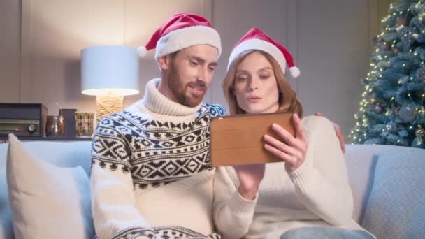 彼女の夫とのかなり美しい女性は,子供や親戚のためのクリスマスや新年のためのタブレットデバイス上の贈り物を遠隔的に選択します. 休日を祝うために準備するサンタクロースの帽子と幸せな家族. - 映像、動画