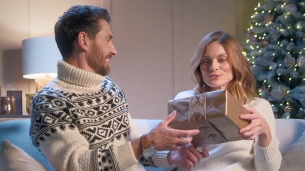 Komea valkoihoinen mies antaa joululahjan rakkaalle vaimolleen. Onnellinen nainen hymyilee iloisesti katsoessaan lahjaa yllätyksenä. Perhe juhlii joulua kotona. Talvilomien käsite. - Materiaali, video