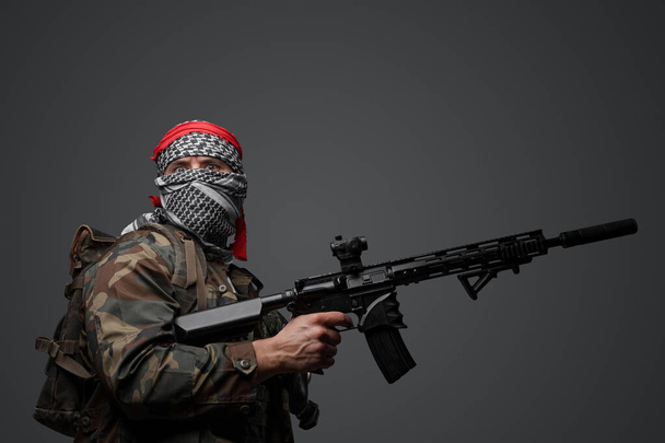 Soldado fanático radical de Oriente Medio vestido con un keffiyeh blanco y un uniforme de campo camuflado, armado con un rifle automático, sobre un fondo gris - Foto, imagen