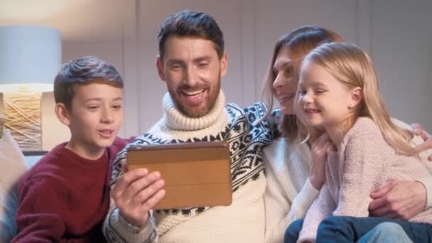 Szerető kaukázusi család pozitív beszélgetést online tabletta eszköz, miközben ülnek együtt kanapén. Gondoskodó apa feleségével és boldog gyermekeivel mosolyog a távoli kommunikáció során. - Felvétel, videó