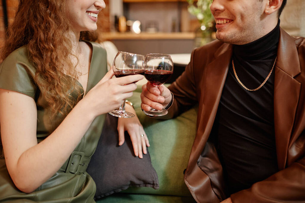 Ευτυχισμένο νεαρό ρομαντικό ζευγάρι σε smart casualwear που κάνει πρόποση με ποτήρια κόκκινο κρασί ενώ κάθεται ο ένας μπροστά στον άλλο στο σπίτι - Φωτογραφία, εικόνα