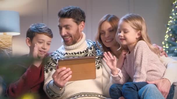 Přátelská bělošská rodina mává rukama během videohovoru na tabletu. Šťastný pár s jejich roztomilé děti mluví online s prarodiči a radostně se usmívá na kameru. Štědrý večer. - Záběry, video