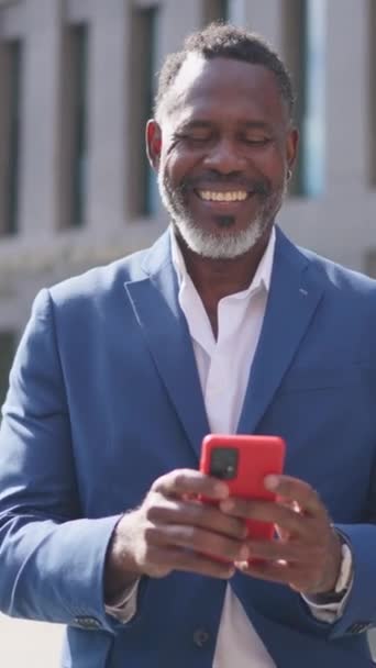 Wideo z zoomem na szczęśliwego afrykańskiego biznesmena korzystającego z telefonu przed budynkiem finansowym - Materiał filmowy, wideo