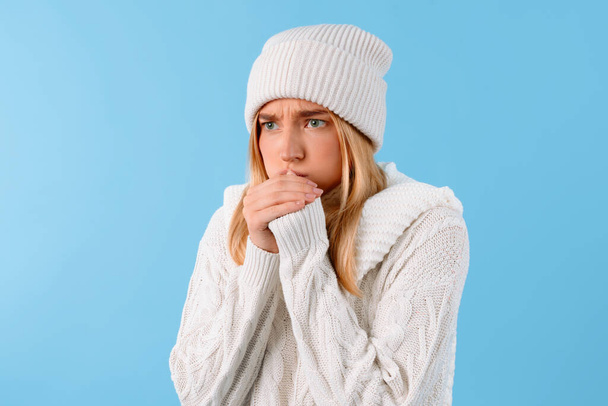 Беспокойная молодая женщина в вязаной шляпе и свитере согревает руки, показывая признаки простуды или гриппа на ярком синем фоне - Фото, изображение