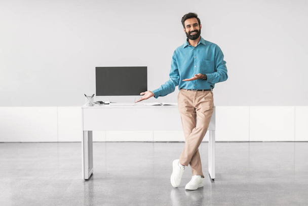 Homme d'affaires indien joyeux geste vers écran d'ordinateur vierge sur bureau blanc, illustrant présentation amicale ou grand site Web d'affaires, maquette - Photo, image