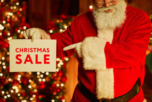 Обрезанный снимок традиционного Санта-Клауса, держащего знак рождественской распродажи и указывающего, стоя в украшенной комнате - Фото, изображение