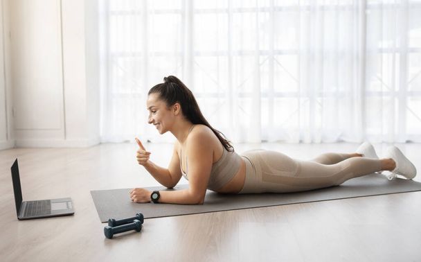 Positive millennial athletic woman have online fitness class, laying on yoga mat, looking at laptop screen and showing thumb up, enjoying her workout on Internet, side view, copy space - Photo, Image