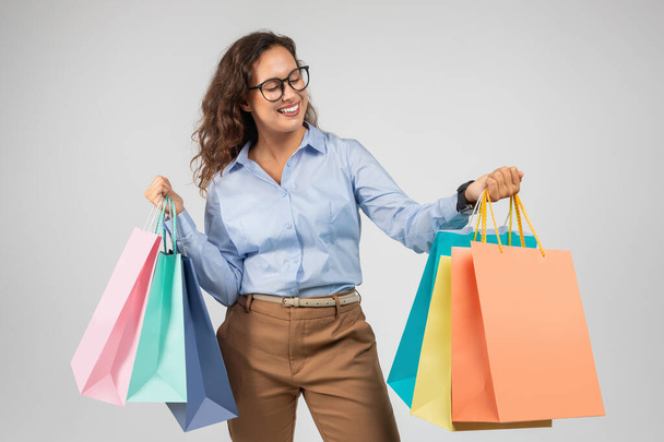 Happy young caucasian lady in formal wear, glasses look at many purchases bags, enjoy shopping, isolated on gray background studio background. Sale, discount, Black Friday - Photo, Image