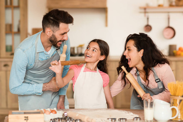 Игривый европейский семейный момент, когда родители и ребенок участвуют в воображаемой битве на мечах со скалками, смеясь вместе в домашней кухне - Фото, изображение