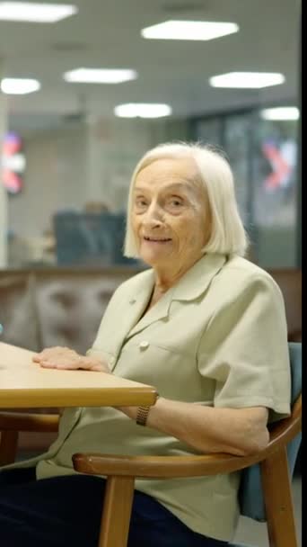 Video vanhasta naisesta katsomassa kameraa istumassa pöydän vieressä pelien kanssa hoitokodissa - Materiaali, video