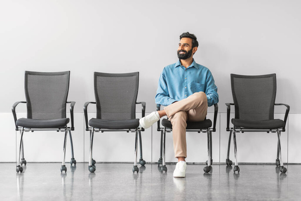 Χαλαρωμένος Ινδιάνος με χαρούμενη συμπεριφορά κάθεται μόνος του σε σειρά από καρέκλες, πιθανόν περιμένοντας συνέντευξη για δουλειά, σε φωτεινό, μοντέρνο χώρο αναμονής - Φωτογραφία, εικόνα