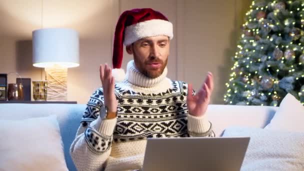 Красивый белый мужчина в шляпе Санта-Клауса разговаривает на видео-вызове на ноутбуке в современной гостиной с рождественской елкой на заднем плане. Удаленно поздравить свою семью или друзей с праздниками. - Кадры, видео