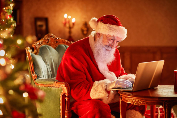 Side view portrait of Santa Claus using laptop by Christmas tree and smiling, copy space - Photo, Image