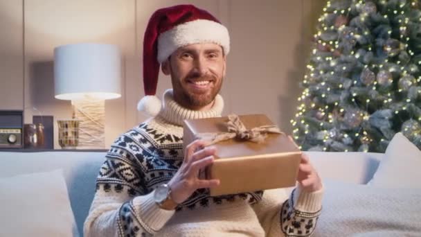 Aantrekkelijke blanke jongeman met kerstmuts vrolijk glimlachend terwijl hij een kerstcadeau vasthoudt. Feestelijke viering thuis. Genieten van het ontvangen van cadeau van vakantie. Traditie. Vrolijk kerstfeest. - Video