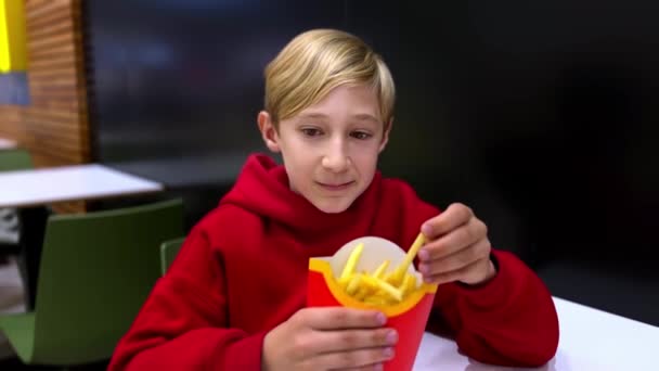 Un chico se sienta en una mesa comiendo papas fritas de una freidora. Un chico en un restaurante comiendo comida rápida - Metraje, vídeo