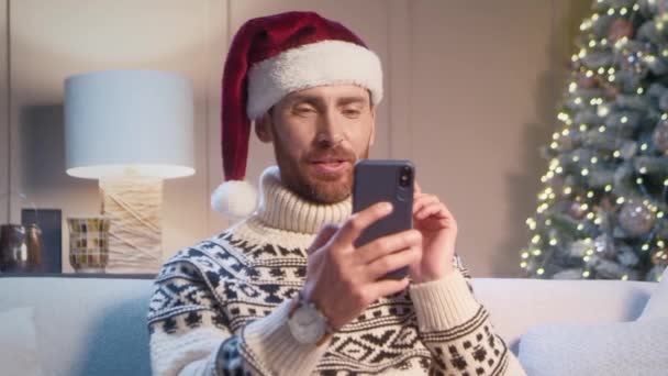 Blanke man met kerstmuts en gebreide trui thuis. Jonge man met communicatie op afstand op zijn telefoon terwijl hij op de bank zit in de buurt van de kerstboom. Begroeting en felicitatie concept. - Video