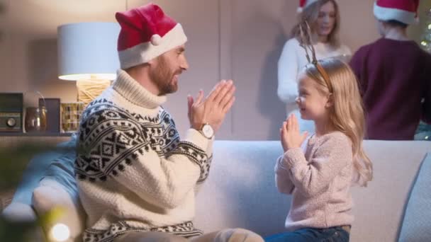 Gondoskodó kaukázusi apa tapsol szerető lányával, miközben anya fiával díszíti a karácsonyfát a háttérben. Boldog család játszik együtt. Az emberek karácsony este pihennek.. - Felvétel, videó