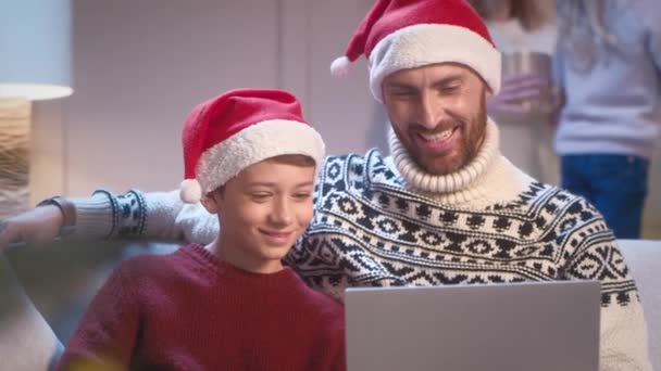 Handsome Caucasian father with his son wearing Santa Claus hats while sitting on sofa in living room. Happy family watching video or funny movie on laptop. Having recreation on Christmas Eve. - Footage, Video