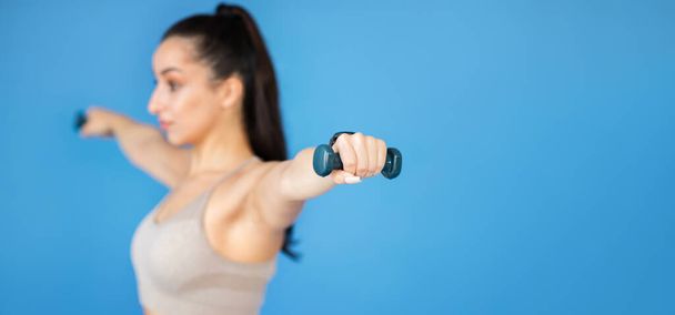Eine fitte junge Frau stemmt Gewichte auf blauem Grund. Shops in Turnbekleidung und Workout, leerer Werberaum neben ihr, Power und Inspiration, Web-Banner - Foto, Bild