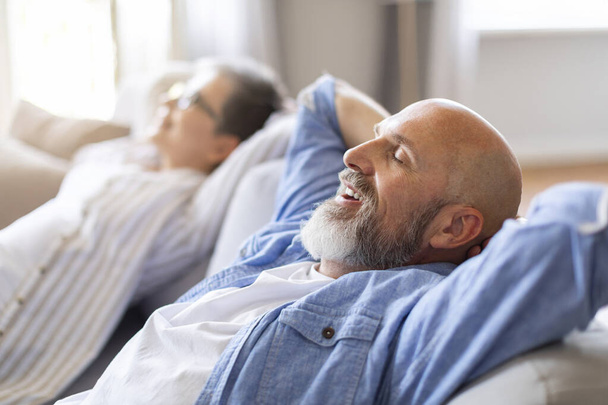 Felices cónyuges mayores relajados apoyados en un sofá cómodo, un hombre sonriente y una mujer descansando con las manos detrás de la cabeza, relajándose en el sofá en la sala de estar, toma de vista lateral con enfoque selectivo - Foto, imagen