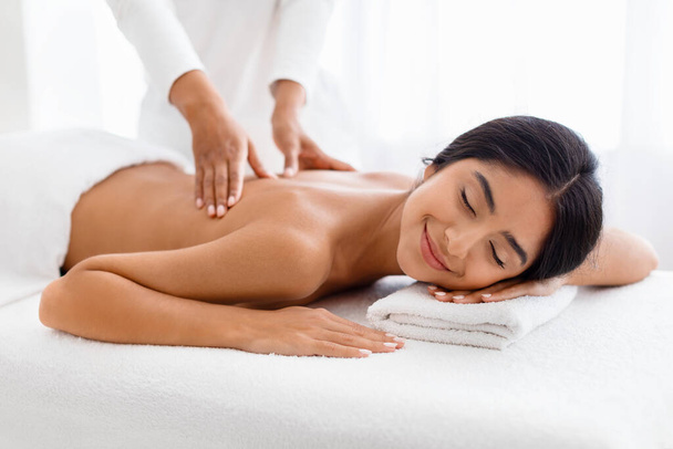 Beautiful Young Indian Woman Enjoying Professional Wellness Massage In Spa Salon, Closeup Shot Of Therapist Lady Massaging Back Of Attractive Client Lying On Table With Closed Eyes - Photo, Image