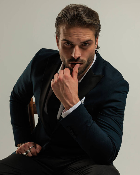 seductor hombre de alta clase en esmoquin mirando hacia adelante y tocando sensualmente los labios mientras está sentado frente al fondo gris - Foto, imagen