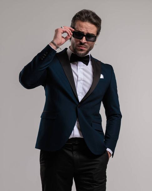 elegant young groom in tuxedo with hand in pocket adjusting sunglasses and posing in front of grey background - Photo, Image