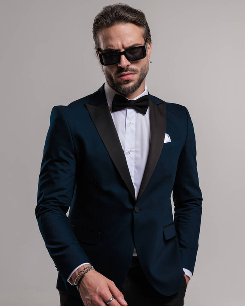 retrato de novio elegante resistente con esmoquin y gafas de sol y posando con la mano en el bolsillo delante de fondo gris  - Foto, Imagen