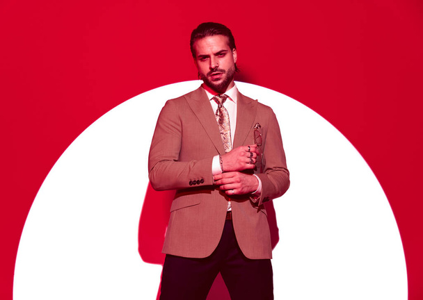 赤い背景のスポットライトの前でシャツの袖を調節しながらスーツに自信を持って優雅なビジネスマン - 写真・画像
