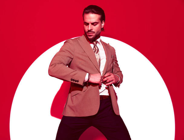 sexy young businessman looking down and closing elegant suit in front of red spotlight background - Foto, afbeelding