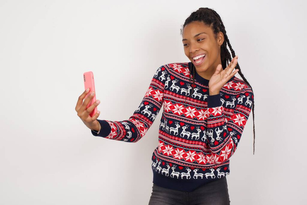 Πορτρέτο της ευτυχισμένης φιλικής Αφρικής Αμερικανός γυναίκα φορώντας χριστουγεννιάτικο πουλόβερ κατά λευκό τοίχο λήψη selfie και κυματίζει το χέρι, επικοινωνία σε βίντεο κλήση, σε απευθείας σύνδεση συνομιλία. - Φωτογραφία, εικόνα