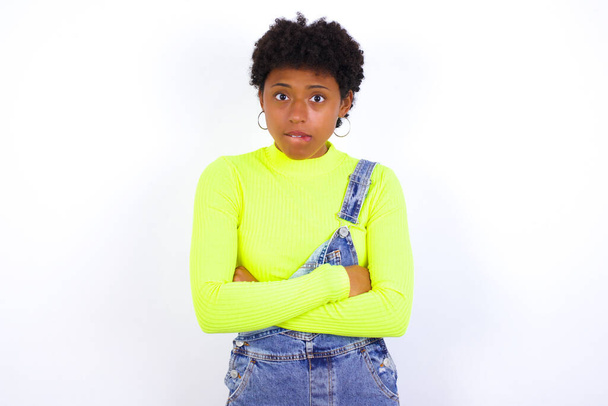 νεαρή Αφρο-Αμερικανίδα γυναίκα με κοντά μαλλιά φορώντας τζιν συνολικά κατά λευκό τοίχο δαγκώνει το στόμα του και αναζητούν ανησυχούν και φοβάται σταυρώνουν τα χέρια, ανησυχία και αμφιβολία. - Φωτογραφία, εικόνα