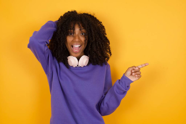 портрет молодої експресивної афроамериканки з навушниками на жовтому тлі, що вказує на порожній простір - Фото, зображення