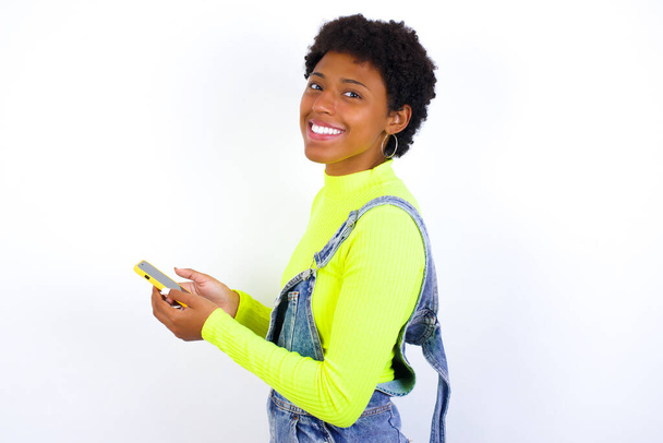 contenu joyeux gai afro-américaine femelle avec bouclée bushy porte jeans salopettes sur mur blanc tenant dans les mains dispositif passe-temps smm post blog - Photo, image