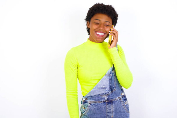 Αστεία νεαρή Αφρο-Αμερικανίδα γυναίκα με κοντά μαλλιά που φοράει τζιν συνολικά ενάντια στο λευκό τοίχο γελάει χαρούμενα, έχει τηλεφωνική συνομιλία, διασκεδάζει από φίλο, κλείνει τα μάτια. - Φωτογραφία, εικόνα