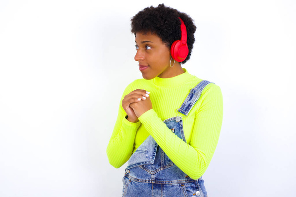 joven mujer afroamericana con el pelo corto con denim en general contra la pared blanca lleva auriculares estéreo escuchando música concentrada y mirando a un lado con interés. - Foto, imagen