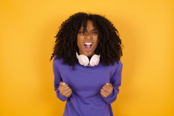 портрет молодої напруженої кричущої афроамериканської жінки з навушниками на жовтому фоні - Фото, зображення