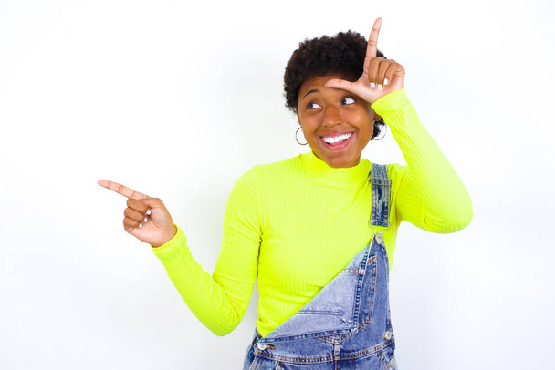 mladá Afroameričanka s krátkými vlasy v džínové kombinéze proti bílé stěně ukazující znamení poraženého a ukazující na prázdné místo - Fotografie, Obrázek