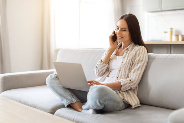 Zdalna kariera. Uśmiechnięta kobieta rozmawiająca przez telefon komórkowy i korzystająca z laptopa w domu, piękna Tysiącletnia kobieta siedząca na kanapie w salonie, pracująca na komputerze i wykonująca telefony komórkowe, wolna przestrzeń - Zdjęcie, obraz