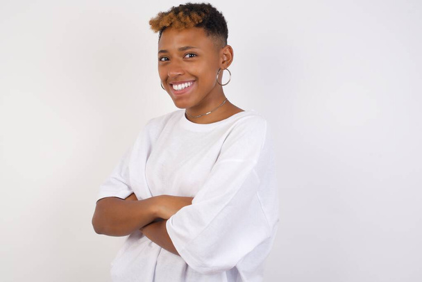Изображение веселой молодой молодой афроамериканской женщины в белой футболке, стоящей в помещении со скрещенными руками. Смотрит и улыбается в камеру. Уверенная девушка. - Фото, изображение